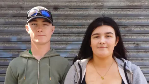 A young man and a young woman are standing together with closed shutters behind them. The man is wearing a cap with sunglasses resting on his head and a green jacket. The woman has long brown hair and is wearing a gold necklace and a grey jacket with a black top.