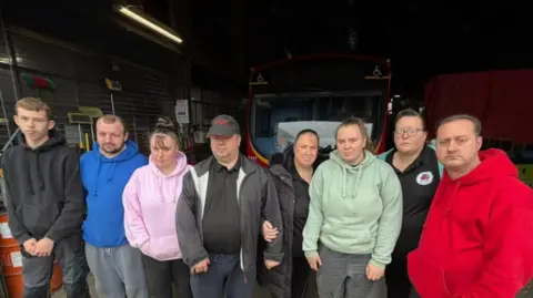 BBC/Jim Scott Volunteers from the Northern National Restoration Group, each with different coloured hoodies, are looking unhappy after preparing to leave their base. In the background is one of their buses, which has a red front and is reflecting sunlight in its windscreen.