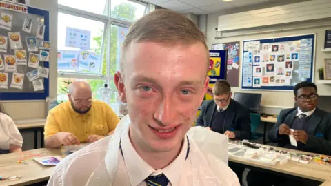 Corey is smiling. He has short fair hair and is wearing a plastic white apron over his school shirt. Pupils are sitting behind desks in the background in a classroom. Decorations are on walls. 