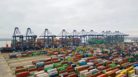 PA A large number of shipping containers sit in front of loaded boats and tall blue machinery. The sea is visible in the background. 