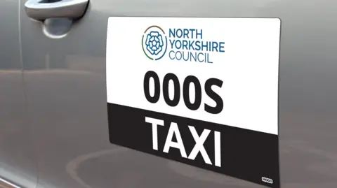 North Yorkshire Council The side of a silver taxi with a sign reading "North Yorkshire Council taxi"