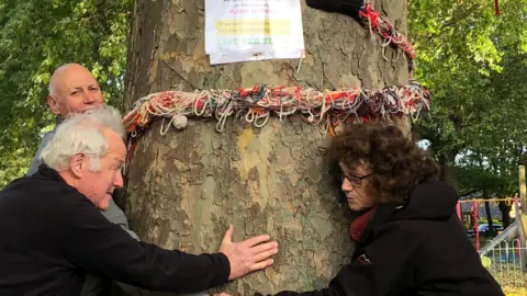 A close up of a large tree with three people showing circling it. They hold hands around the tree . A multi coloured rope is also around the tree with a laminated sign saying 'save out tree' 