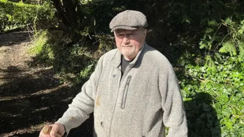 BBC/Phil Harrison An elderly man with a flat cap and a grey fleece looks at the camera. It is a sunny day. He carries a stick. He is standing outside shaded by trees.