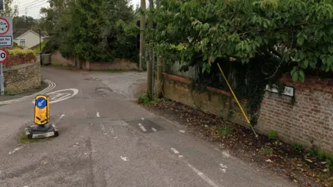 Google Maps One entry to Dark lane, with the road sign showing on the right of the picture. There is a bollard in the road and an accept for access street sign to the left. There are trees in the picture, around, and above, walls as you enter the road. 