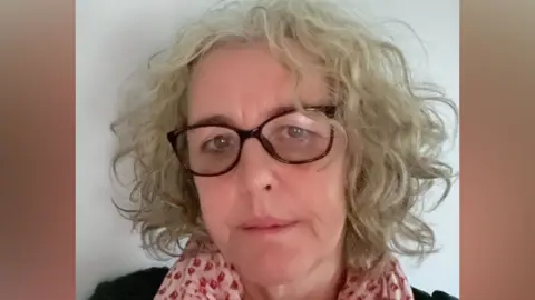Nicki Durbin in a selfie-style image. She is slightly smiling at the camera. She has short grey curly hair and wears glasses along with a pink scarf and black top.
