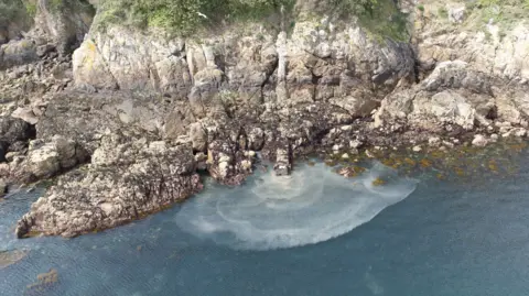 A cliff face with a small under the rock pipe coming down it. From the pipe a brown water (sewage) is being released, slowly coming out in a semi-circular pattern to the blue sea. 