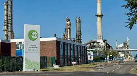 The entrance to an oil refinery with a large white and green sign, reading "Prax Lindsey Oil Refinery Main Entrance" in front of a two-storey red-brick and glass building. A road leads towards tall metallic chimneys and other refinery structures under a blue sky.