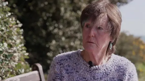 Angela Hardy, a woman with short dark hair, is pictured on a bench in front of some bushes. She has an earring dangling from her left ear and is wearing a blue woolly jumper. 