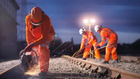 Getty Images Three railway workers in orange hi-vis overalls working on railway track under floodlights. They are all wearing hard hats. One has a power tool which is creating sparks as it hits the metal of the permanent way. The others have tools which they are applying to the track.