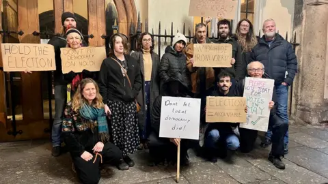 About a dozen people standing outside the Guildhall in Exeter with some holding signs written on cardboard and calling for elections to go ahead in May.
