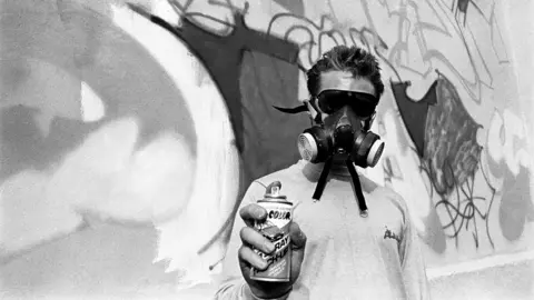Beezer Picture taken by Beezer, of a man in a mask doing 3D graffiti art spray in St Paul's, Bristol, in 1985. The man is holding the spray can towards the camera with the graffiti wall behind him.
