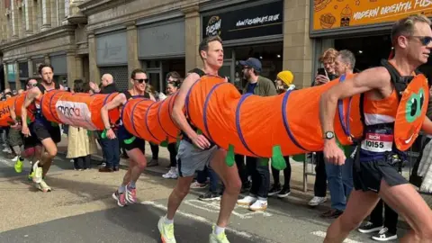 MND Association Men running in an orange caterpillar costume. They are joined at the chest and back by a concertina-type fabric.