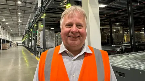 A man with a mop of blonde hair smiles as he stands in a large warehouse-type cold storage facility with rows of low and high shelves behind him. He is wearing an orange high-vis vest over a grey shirt.