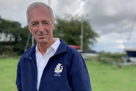 Man in  a navy blue zip up jacket is looking at the camera with boats in the background behind him