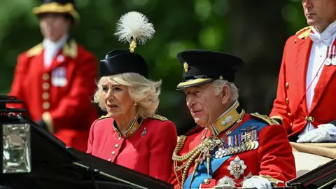 Getty Images The King and Queen riding in a carriage. He is wearing the uniform of the Grenadier Guards as well as a number of medals. Queen Camilla is wearing a red, silk crepe, grenadier guards uniform dress and a black beret.