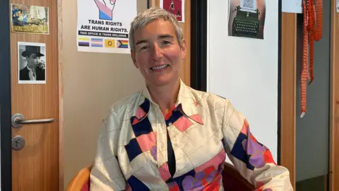 A woman with short grey hair and a cream-coloured blouse with some colourful patches sits in a chair, smiling at the camera. The wall behind her is decorated with pictures and posters, including one which reads: "TRANS RIGHTS ARE HUMAN RIGHTS".
