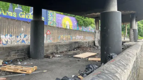 Concrete columns hold up a flyover type road. a number of wooden pallets and other debris can be seen in the space under the road