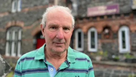 Richard Edwards, looking at the camera. He has thin grey hair, and is wearing a green and blue striped polo shirt. He is stood outside his guesthouse in Betws-y-Coed