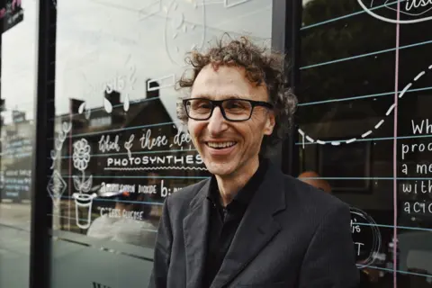 BBC/Caroline Briggs Andrew Gough wearing a grey jacket and black shirt and glasses stands in front of a window covered in white writing and doodles. 