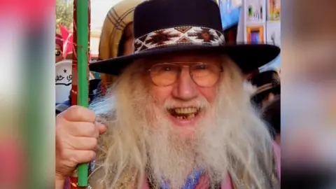 John Row. He has long white hair and a long white beard. He is wearing glasses and a black hat. He is looking at the camera and smiling with his mouth open slightly.