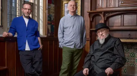 Royal Shakespeare Company A man with short hair and a beard in a blue cardigan standing next to a man with a bald head and pale blue shirt and a seated man with a white beard, black shirt and black hat