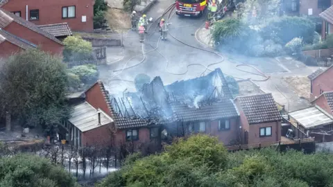 The destroyed roofs of bungalows can been seen, with smoke rising from them. There are other houses in a small road and a fire engine and firefighters are nearby