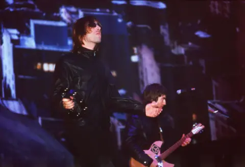 Getty Images A long haired man with sunglasses and wearing a black leather jacket is holding a tambourine while another long haired man behind him plays a guitar