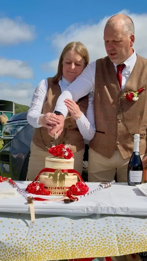 Family photo Safron and Nigel stood together cutting their cake on a table in front of them. The cake is cream with red flowers on and stands next to a bottle of prosecco. 