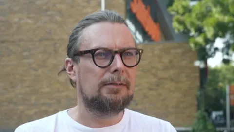 Close-up portrait of Matt Arnold, a man with light brown hair tied back, glasses, and a beard, wearing a white t-shirt outside with a Sainsbury's store blurred in the background.