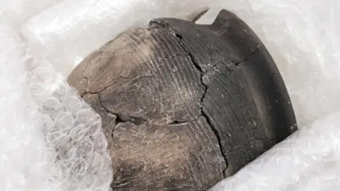 East West Rail A fragile ceramic vessel is shown carefully wrapped in protective bubble wrap for storage or conservation. The dark-coloured pot displays visible cracks and repaired sections, indicating its age and delicate condition. Fine ridged decoration can be seen on the surface, highlighting details of its craftsmanship. The image suggests an archaeological artefact being preserved or prepared for examination and display.