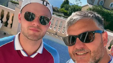 BBC Two men wearing sunglasses are looking at the camera. One is wearing a Villa shirt with a white cap on backwards and stubble. The other has a beard and grey hair. The sun is shining and there is greenery behind.