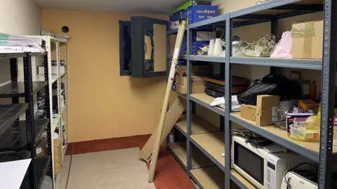 Edd Smith/BBC A room filled with metal shelving on which there are various items, including a microwave and lights. On the back wall is a large square metal escape hatch. 