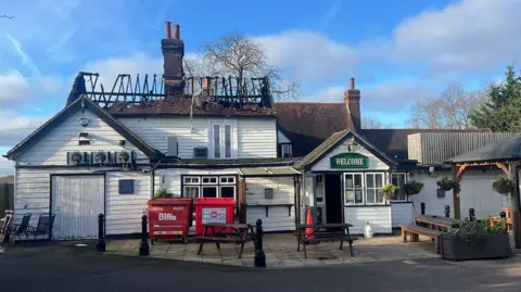 Richard Knights/BBC The gutted roof of the pub the following morning. A few smoke-blackened beams are all that remain of a large section of roof. Red bins still stand outside the pub and there are tables and benches outside too. The pub walls is painted white.