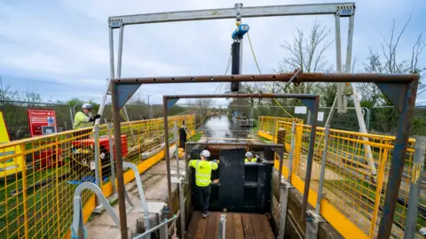 A lock in a canal, with workers in hi-vis jackets working on it and yellow fencing on either side.