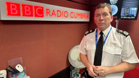 BBC Library image of Rob Carden, chief constable of Cumbria Police, pictured in one of BBC Radio Cumbria's studios. He is wearing his police uniform - a white shirt with black tie. On the wall to his right-hand side is a large BBC Radio Cumbria sign.