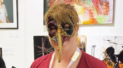Rachael Bassett A woman wearing a Venetian mask at an art exhibition. The mask is black and gold and has a long nose section that looks like a beak. She wears a red T-shirt and lanyard. Pieces of art are on display behind her.