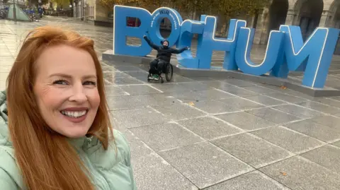 Anna Thomas Selfie of a woman with long red hair wearing a turquoise down jacket and smiling at the camera. Behind her are large blue letters spelling out BOCHUM, and in front of her is a man dressed all in black, sitting in a wheelchair with his arms raised.