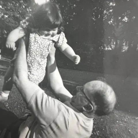 Marcia Lewis A black and white photo of Anthony Pratt holding his daughter in the air as a young girl. He is lying on the grass, with trees in the background