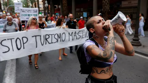 Reuters A demonstrator with tattoos uses a megaphone as people protest against mass tourism on a street in Alicante, Spain, July 13, 2024. The banner reads 