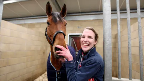 Sophie Christiansen with horse in stable is smiling at the camera and wearing a navy blue fleece.