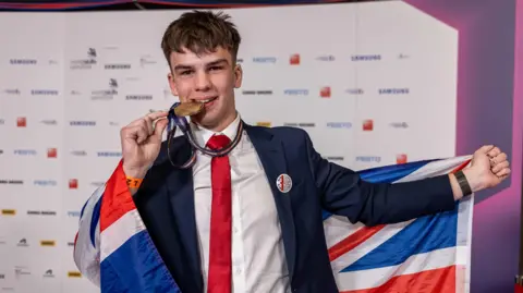 WorldSkills UK A man dressed in a navy suit with a white shirt and red tie. He is pictured holding and pretending to bite a bronze medal whilst holding a Union Jack flag.