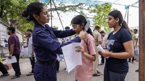 A female security woman is running a hand-held metal detector over a young woman's hair, as others wait in line.