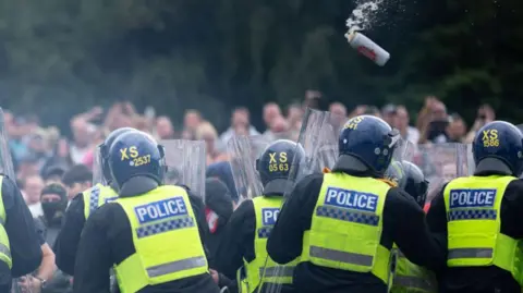 Getty Images Point of view is standing behind police officers holding riot shields. A beer can is about to hit one of them. In the distance there are many blurred figures taking part in the "protest".