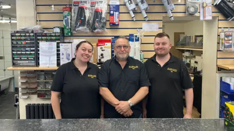 Three people stand in a row behind a desk in a plumbing supplies shop. A woman stands to the left of an older man, with a younger man on his right. They all wear black t-shirts, emblazoned with a yellow logo on the right. Behind them on the wall is a selection of plumbing accessories. They are smiling for the camera.