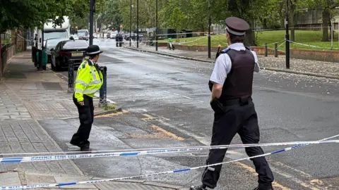 PA Media Two police officers stand by the side of a section of the road which has been cordoned off