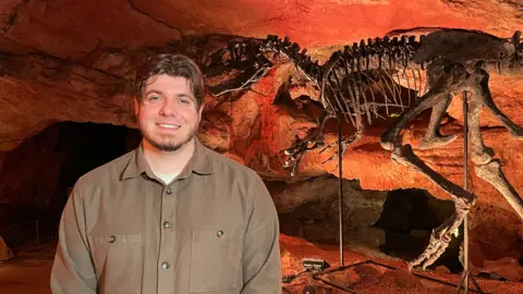A man in a khaki shirt smiles at the camera. He has brown hair and a small beard. Behind him stands the skeleton of an Allosaurus.
