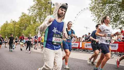 A man dressed in a polar bear onesie and wearing a blue bib, and looking exhausted, raises his right fist up partially while running in the London Marathon.
