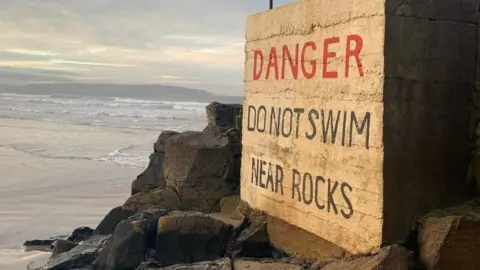 Portstewart Strand. A large sign reads: 'Danger – Do Not Swim Near Rocks.' It is positioned beside a wide area of jagged black rocks, with the shoreline visible in the background