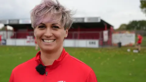 Woman with light pink hair is smiling. She is wearing a red football shirt with the logo Downham Town FC in white.