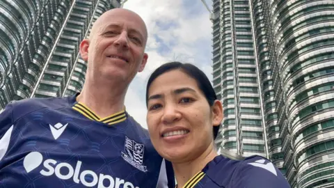 A man and a woman standing next to each other wearing Southend football shirts. Behind them are skyscrapers.